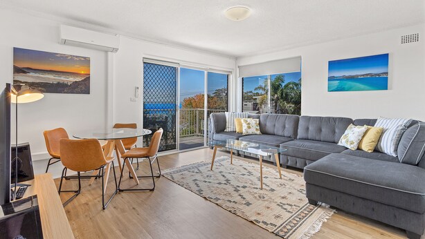 A modern living room with a glass table, grey sofa, and two chairs. There are large windows with a view of the outdoors. - Yarramundi a Nelson Bay Apartment Near Beach (Nelson Bay)