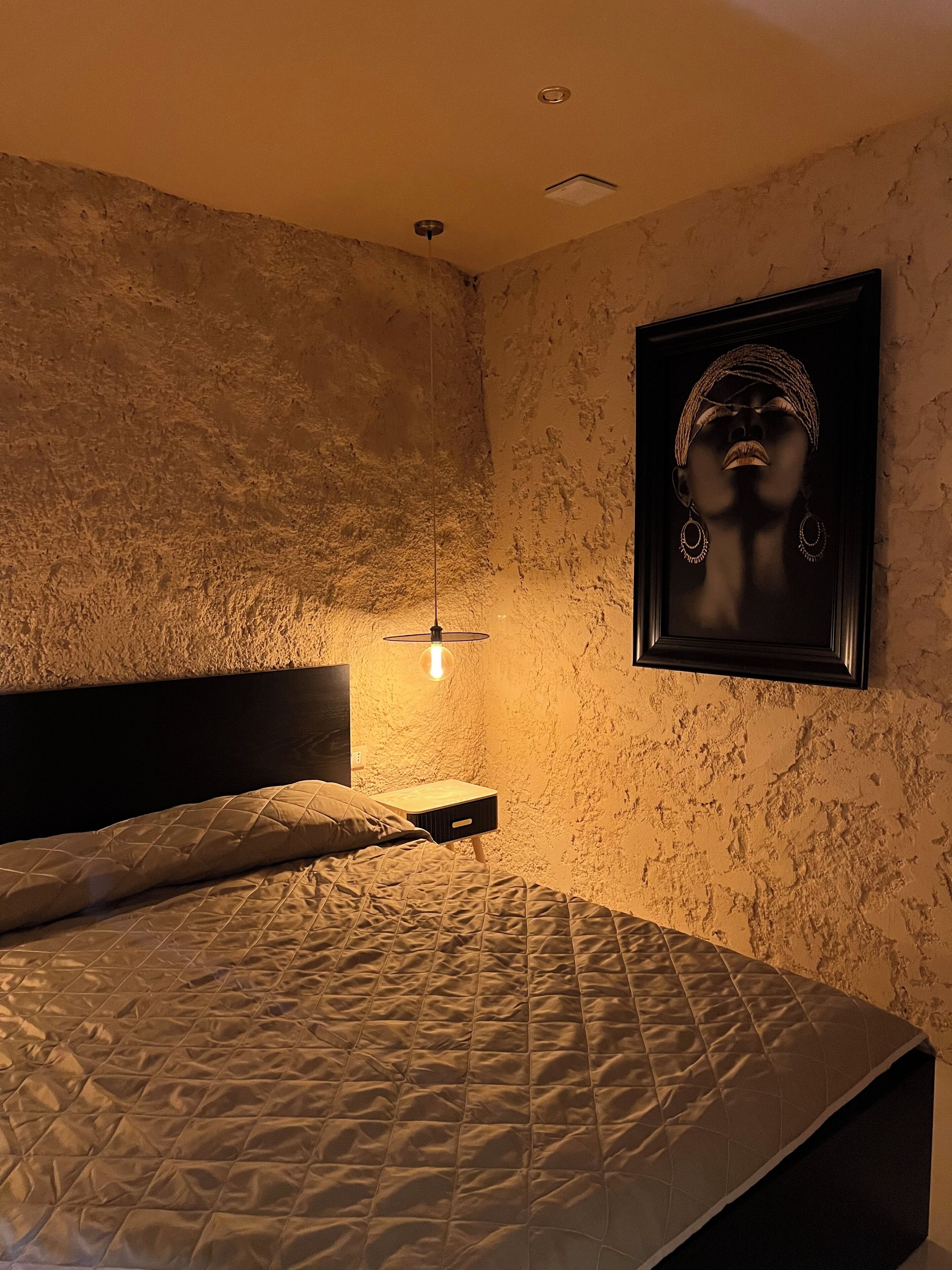 Een slaapkamer met een structuurwand, een bed met een gewatteerde spreidek, een nachtkastje met een lamp, en een lijst met een foto aan de muur.