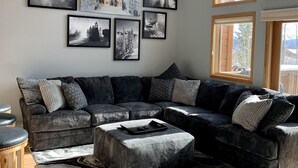 Ein modernes Wohnzimmer mit einem Ecksofa, einem Couchtisch und einem großen Teppich. An der Wand befinden sich mehrere gerahmte Fotografien, und das Zimmer verfügt über große Fenster mit Holzrahmen.