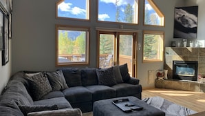 Ein modernes Wohnzimmer mit einer Eckcouch, einem Kamin und großen Fenstern, die auf eine Terrasse und einen Wald Blick bieten.