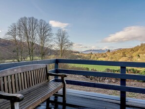 Eine hölzerne Bank mit Metallgeländer, die über eine grüne Landschaft mit Bäumen und Hügeln blickt.