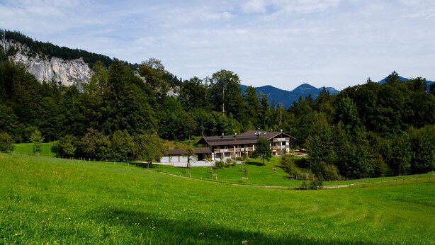 Una casa con techo inclinado, rodeada por un campo verde y árboles, con montañas al fondo.