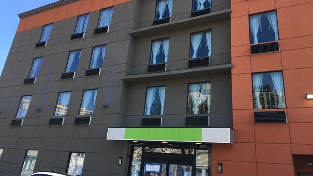A multi-story hotel with a modern facade, featuring a mix of orange and grey panels, large windows with curtains, and a green sign above the entrance. - Magna Hotel (Jamaica)