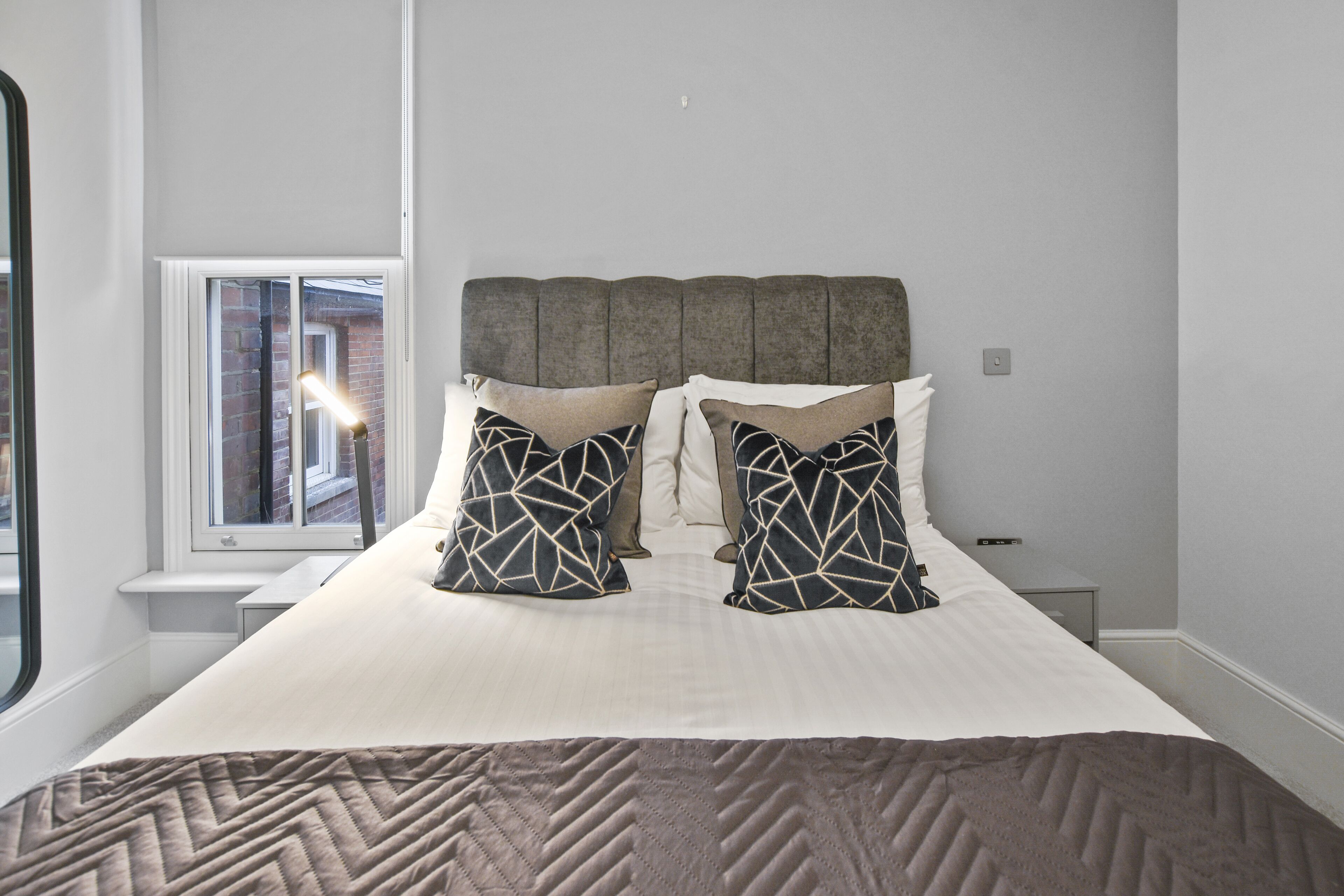 A neatly made bed with a geometric patterned pillow, a headboard, and a window with blinds.