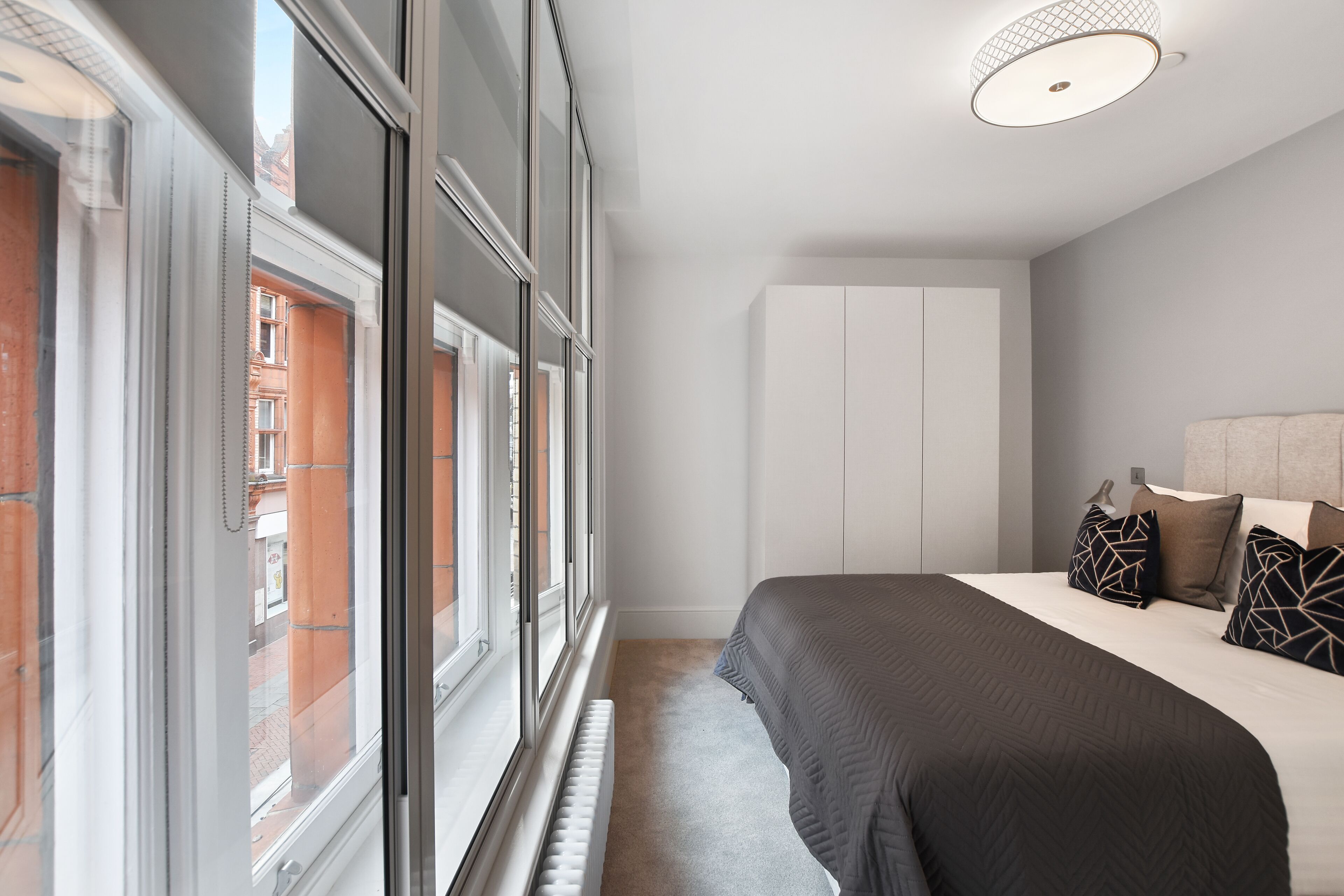 Une chambre avec une grande fenêtre, un lit recouvert d’une couverture sombre et une armoire blanche.