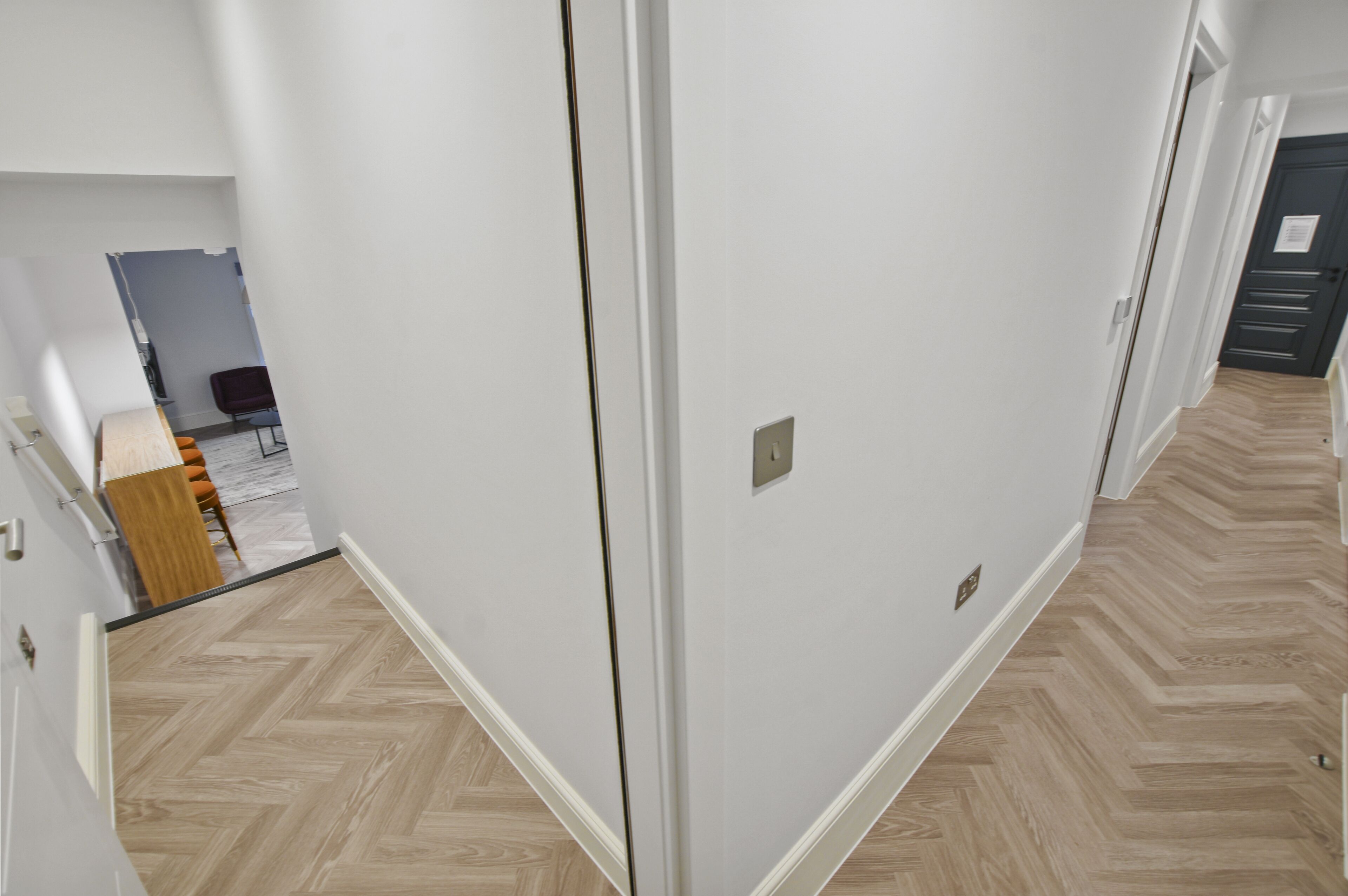 A hallway with a herringbone floor pattern, white walls, and a door leading to another room.