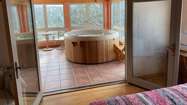 A room with a wooden hot tub, a small table, and chairs, with large windows offering a view of trees. - Luxury Million Dollar Mountain Views: Golf Course Community across Ski Area!  (Angel Fire)