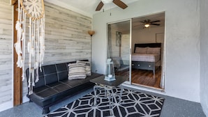 Un salon moderne avec un canapé en cuir noir, un tapis à motifs géométriques, un attrape-rêves, un ventilateur de plafond et une porte en verre donnant sur une autre pièce.