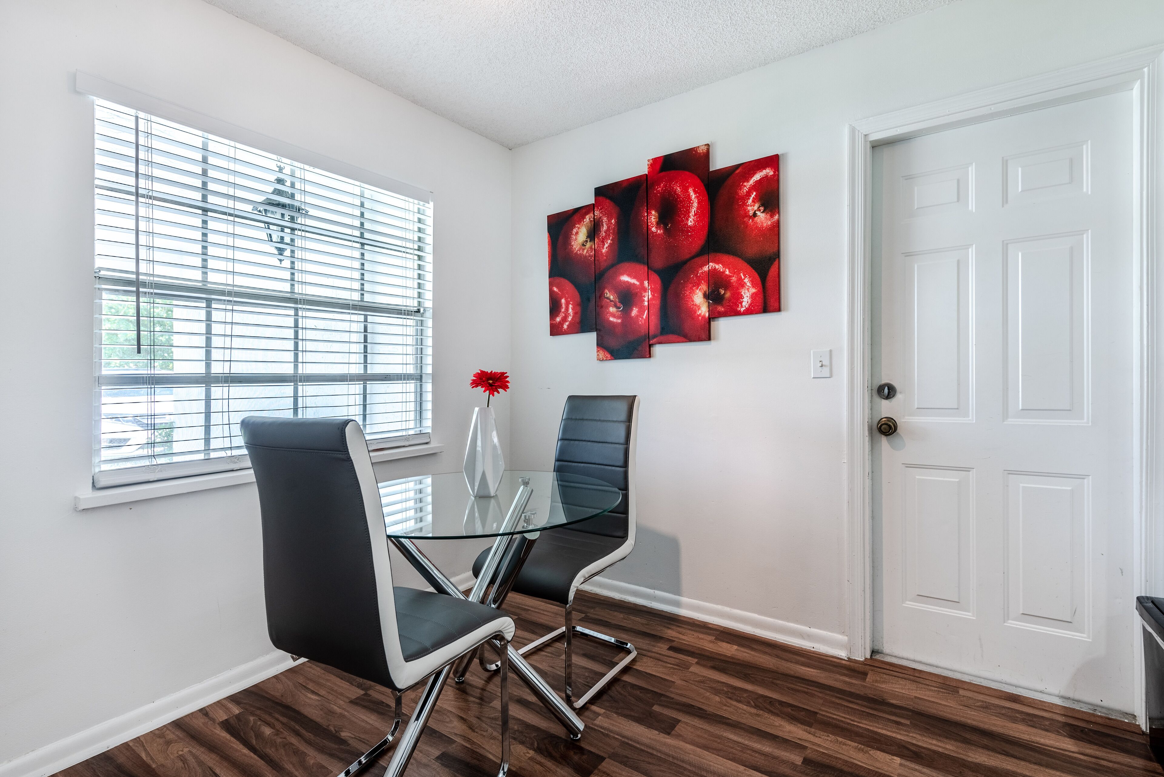 A modern meeting room with a glass table, two black chairs, a red apple painting, and a white door.