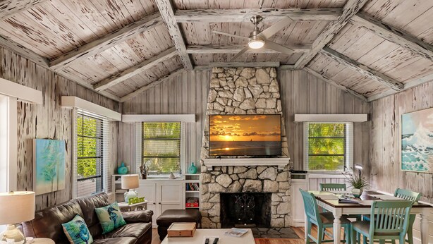 A cozy cabin interior with a stone fireplace, wooden ceiling beams, a leather sofa, and a dining area with teal chairs. - Vintage Cottage from 1936 across street from Beach  and  Heated Pool (Siesta Key)