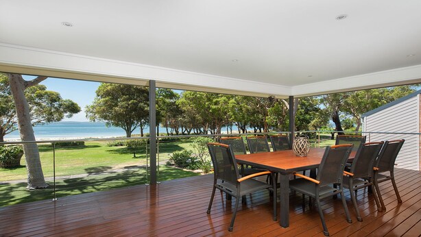 A covered outdoor dining area with a wooden table and chairs, overlooking a grassy landscape and the sea.
