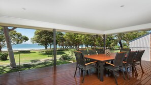 A covered outdoor dining area with a wooden table and chairs, overlooking a grassy landscape and the sea. - Memories on the Beachfront (Corlette)