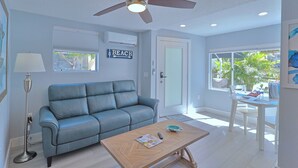 A living room with a blue sofa, a wooden coffee table, a ceiling fan, and a window with a 'BEACH' sign. - Nook Cottage Across street from beach  and  Heated Pool (Siesta Key)