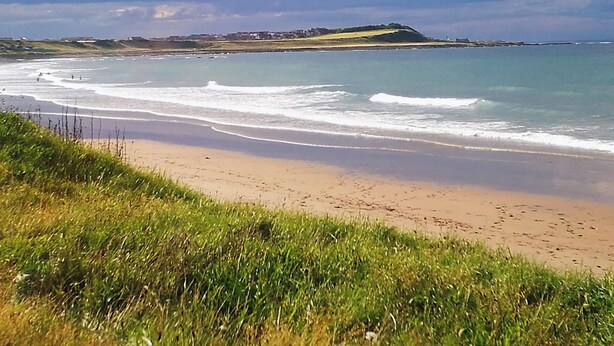Une plage avec des vagues, des zones herbeuses et, au loin, une colline parsemée de bâtiments.