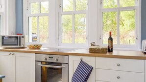 Une cuisine avec des meubles blancs, un four à micro-ondes, un four traditionnel, une fenêtre donnant sur des arbres et une bouteille de vin posée sur le plan de travail.