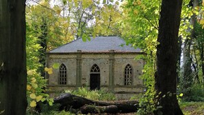 Un bâtiment rustique en pierre, doté d’un toit en pente, entouré d’une végétation luxuriante et avec un arbre abattu en premier plan.