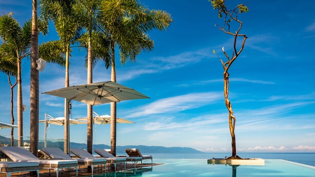 Ein Infinity-Pool mit Liegestühlen und Sonnenschirmen, umgeben von Palmen und blauem Himmel.