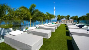 Reihenweise Betten mit weißen Laken und Kissen, auf einer Rasenfläche mit freiem Blick auf das Meer und Palmen.