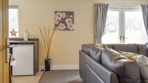 A living room with a grey sofa, a small white fridge, a vase with branches, and a floral painting on the wall.