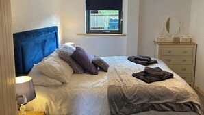 Een netjes opgemaakt bed met een blauw hoofdeinde, een bedlampje en een raam met uitzicht op een achtertuin.