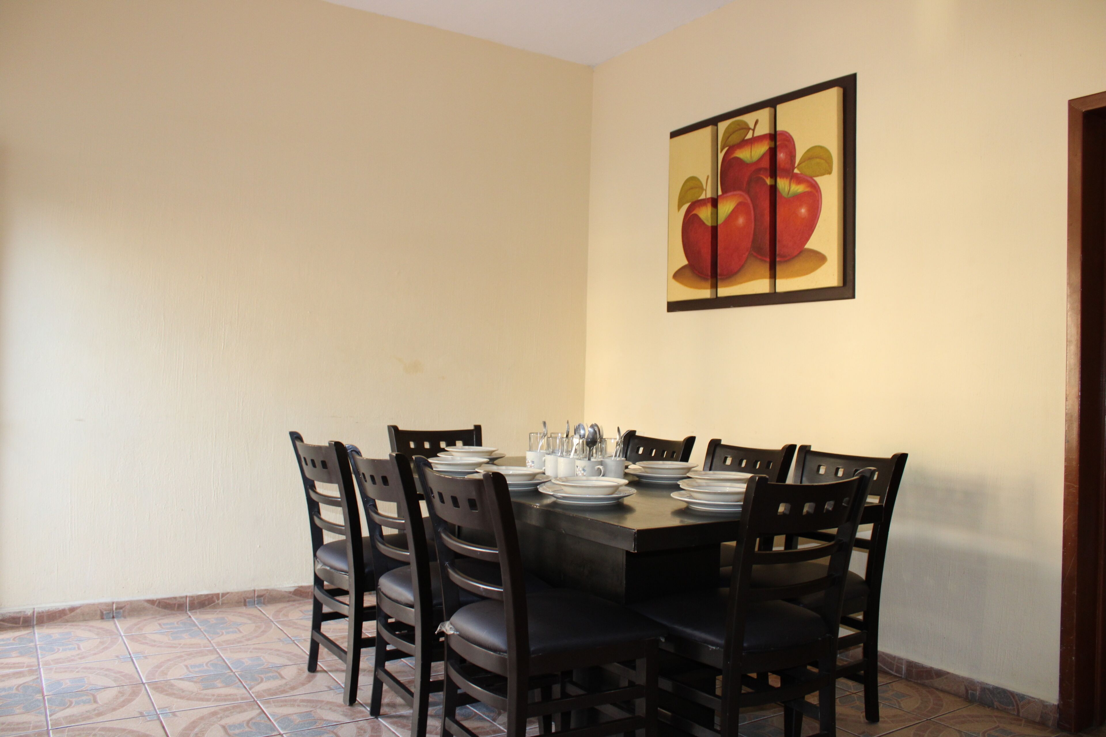 Uma sala de jantar com uma mesa posta para quatro pessoas, rodeada por cadeiras, e um quadro com a imagem de maçãs na parede.