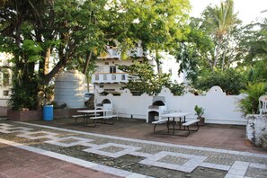 An outdoor patio with picnic tables and a barbecue area, surrounded by greenery and a white wall. - Bungalows Yessy Tetos (Rincón de Guayabitos)