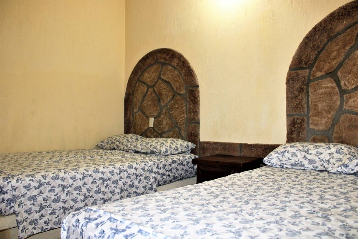 A room with two beds, each with a floral-patterned bedspread and matching pillows. The beds have stone headboards and are positioned against a plain wall.