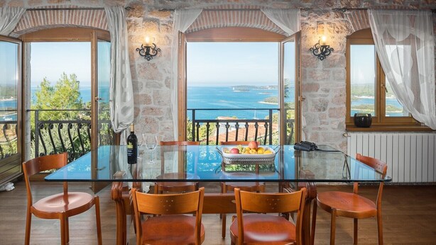 A dining area with a glass table and wooden chairs, overlooking a scenic view of the sea and greenery. - Stunning stone house nestled in the heart of the historic old town of Vrsar. (Vrsar)