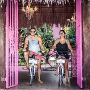 Two people on bicycles in a doorway with pink railings and a thatched roof.