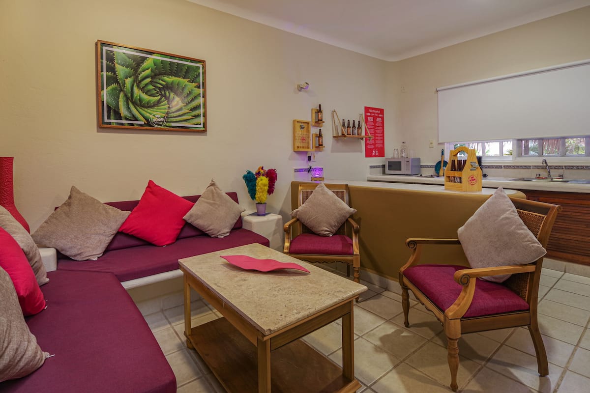 Een woonkamer met een bank, stoelen, een salontafel en een keukenhoek met een spoelbak en magnetron.