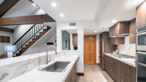 A modern kitchen with marble countertops, wooden cabinets, stainless steel appliances, and a staircase leading to another level.