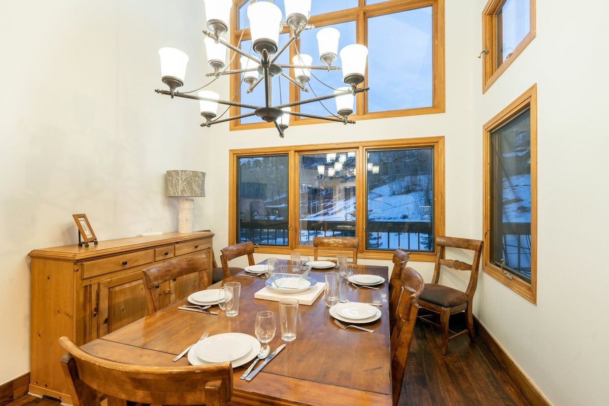 Een eetkamer met een houten tafel gedekt voor zes personen, een kroonluchter en grote ramen met uitzicht op een besneeerd landschap.