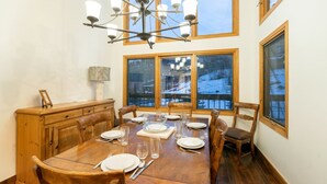 A dining room with a wooden table set for six, a chandelier, and large windows with a view of a snowy landscape. - Kayenta II 16 3 Bedroom/3 Bathroom Slopeside (Mountain Village)