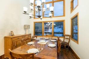 Une salle à manger avec une table en bois dressée pour six personnes, un lustre et de grandes fenêtres donnant sur un paysage enneigé.