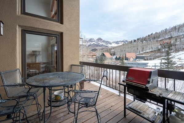 A deck with a round table and chairs, a grill, and a view of a snowy mountain range.