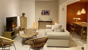 Un salon chaleureux avec un canapé, deux tables rondes, des fauteuils en osier, un téléviseur, un vase décoratif et un tableau accroché au mur.