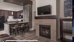 A modern kitchen with dark cabinets, a central island with bar stools, a built-in microwave, and a flat-screen TV above the fireplace.