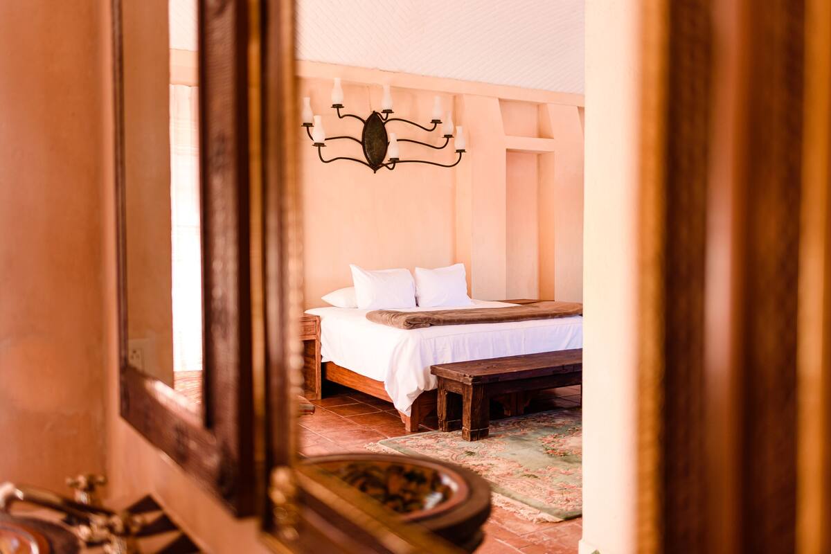 A bedroom with a bed, a bench, a wall-mounted light fixture, and a mirror.