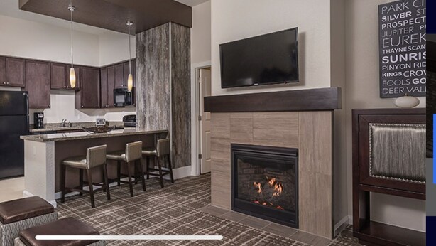 A modern kitchen with dark cabinets, a central island with bar stools, a built-in microwave, and a flat-screen TV above the fireplace.