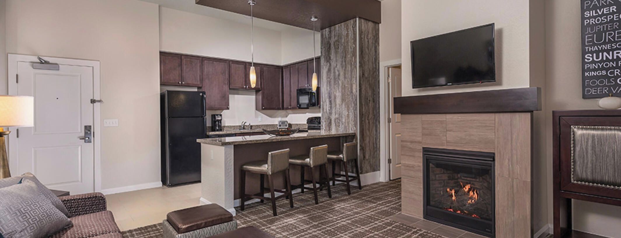 A modern kitchen with dark cabinets, a central island with bar stools, a built-in microwave, and a flat-screen TV above the fireplace.