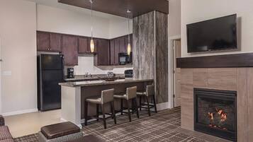 A modern kitchen with dark cabinets, a central island with bar stools, a built-in microwave, and a flat-screen TV above the fireplace.