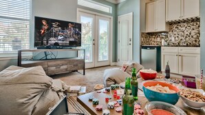 Un salon avec un téléviseur diffusant un concert, une table basse avec des collations et des boissons, et un coin cuisine avec des placards et un pare-feu en carreaux.