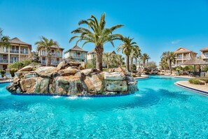Une piscine d’un complexe hôtelier, avec un îlot rocheux, des palmiers et plusieurs bâtiments d’hôtel en arrière-plan.