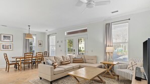 Un salon spacieux avec un canapé d’angle beige, une table basse en bois, un coin repas comprenant une table et des chaises en bois, et un téléviseur posé sur un meuble en bois.