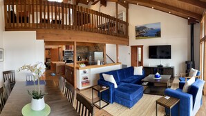 A spacious living area with a high ceiling, wooden beams, a mezzanine level, a kitchen with modern appliances, and a large TV.