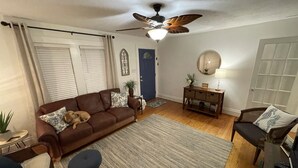 Ein Wohnzimmer mit braunem Sofa, zwei blauen Sesseln, einem Holztisch, einem Deckenventilator, einer Holzkonsole und einer Tür mit blauem Rahmen.