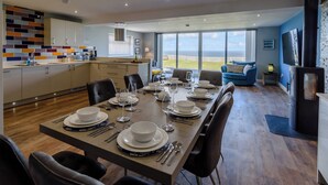 Un espace repas moderne avec une grande table dressée pour six personnes, une cuisine agrémentée d’un mur de crédence aux couleurs vives et une vue sur la mer grâce à de grandes fenêtres.