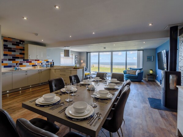 A modern dining area with a large table set for six, a kitchen with a colorful backsplash, and a view of the sea through large windows. - 4 bedroom accommodation in Reighton Gap, near Filey (Reighton Gap, near Filey)