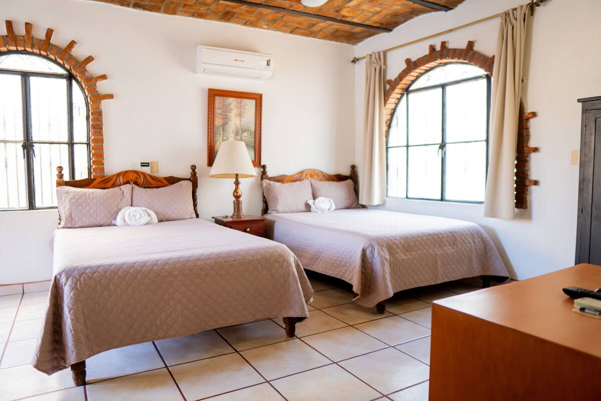 Habitación de hotel con dos camas, un escritorio y ventanas grandes con arcos.