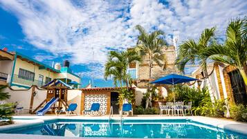 A swimming pool with lounge chairs and umbrellas, surrounded by palm trees and buildings with a clear blue sky.